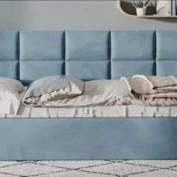 Łóżko z tapicerowanym, błękitnym zagłówkiem w kwadraty, pościelone białą pościelą i beżową narzutą, na podłodze wzorzysty dywan.