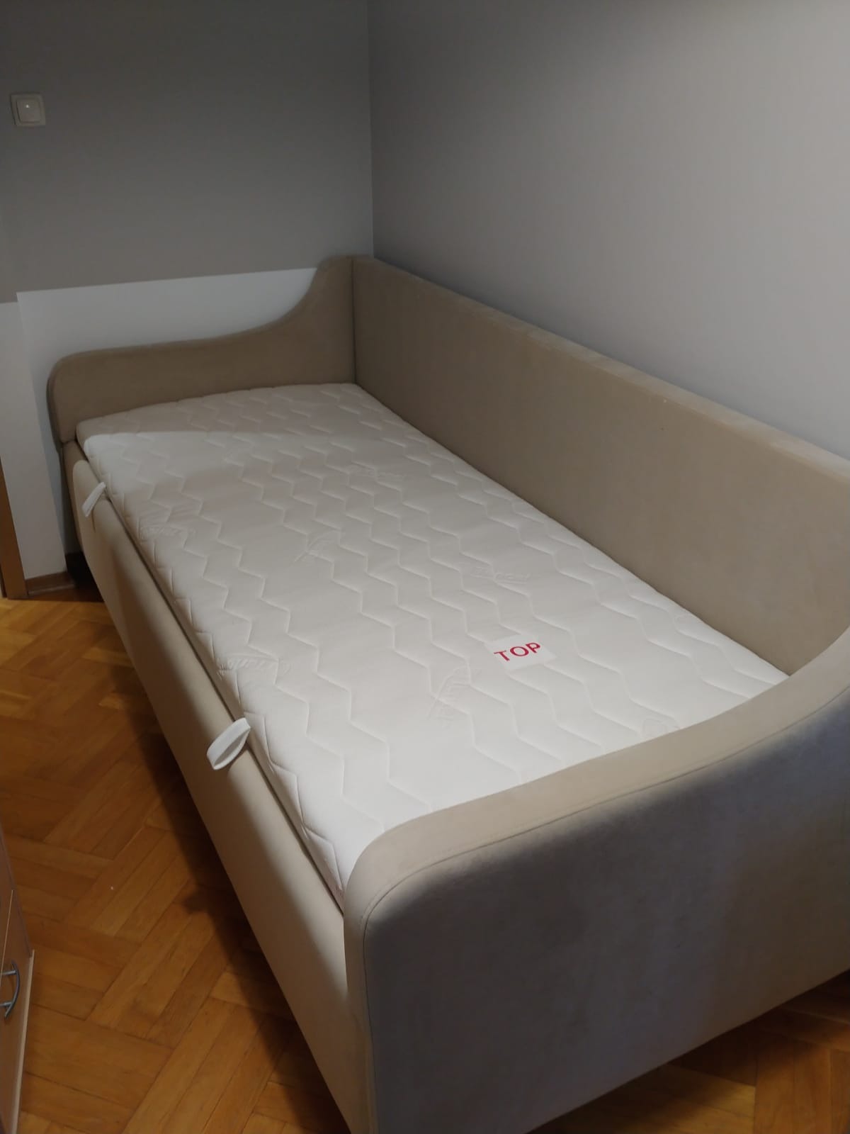 Tapczan z beżową tapicerką i materacem ze wzorem w romby, widoczny w rogu pokoju na parkiecie.