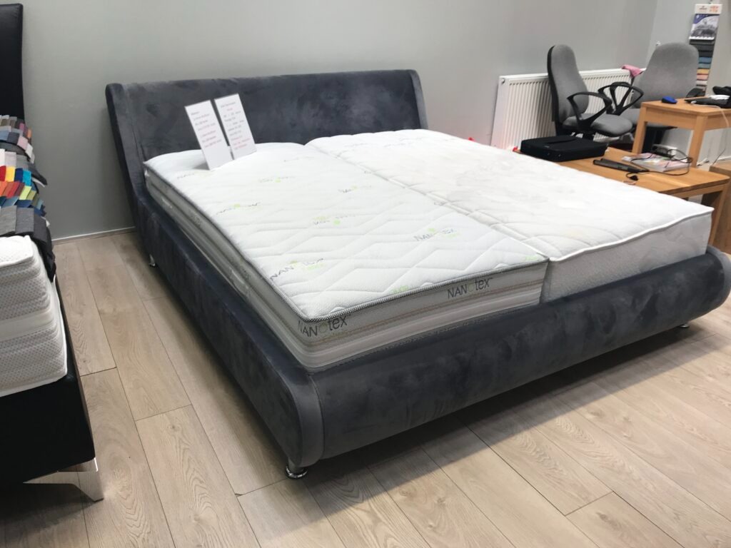 Szare, tapicerowane łóżko z dwoma materacami o różnym stopniu zużycia, w tle biuro z krzesłami.