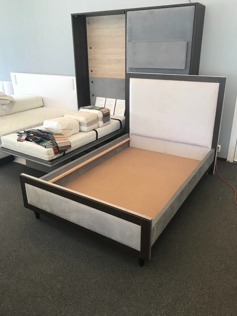 Rama łóżka tapicerowanego z wezgłowiem, w szarym obiciu i z ciemnobrązowymi elementami wykończeniowymi, na tle ekspozycji meblowej z próbkami materiałów.