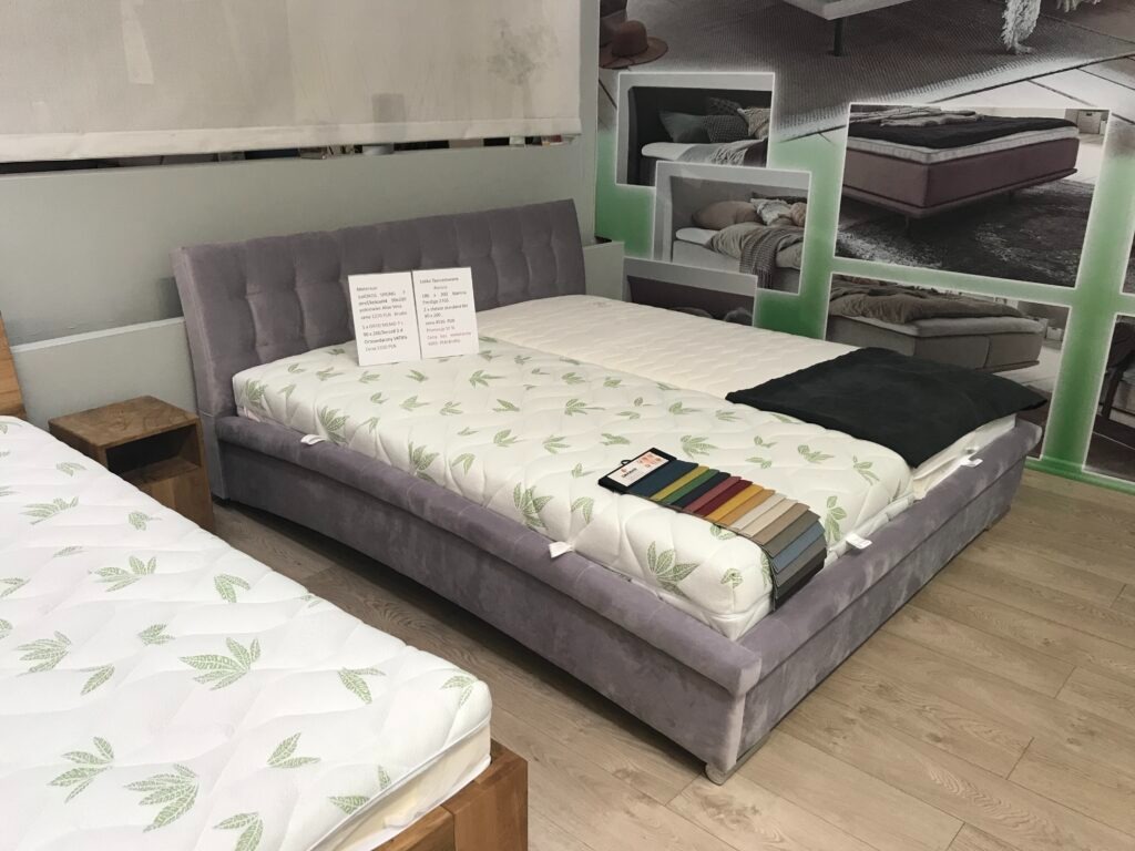 Szare łóżko tapicerowane z zagłówkiem, materacem w liście, próbkami tkanin i czarną poduszką, prezentowane w sklepie obok innego łóżka z podobnym materacem.