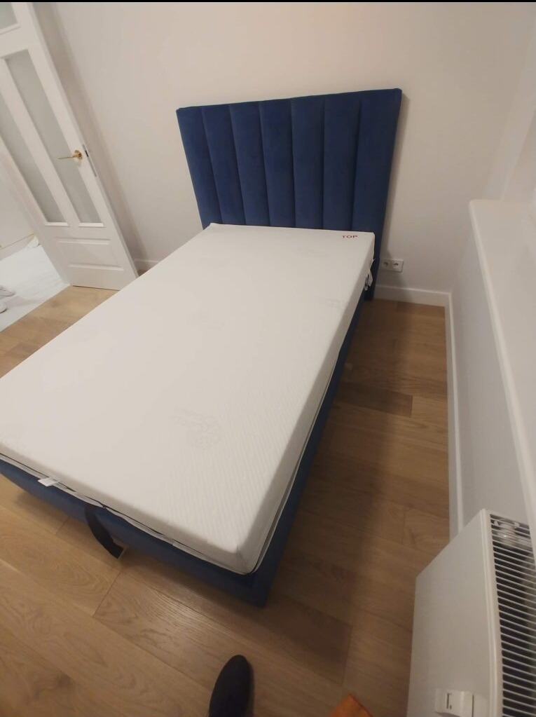 Niebieskie łóżko tapicerowane z wezgłowiem w pionowe pasy, biały materac, drewniana podłoga, widok z boku z lekkiego ukosa.