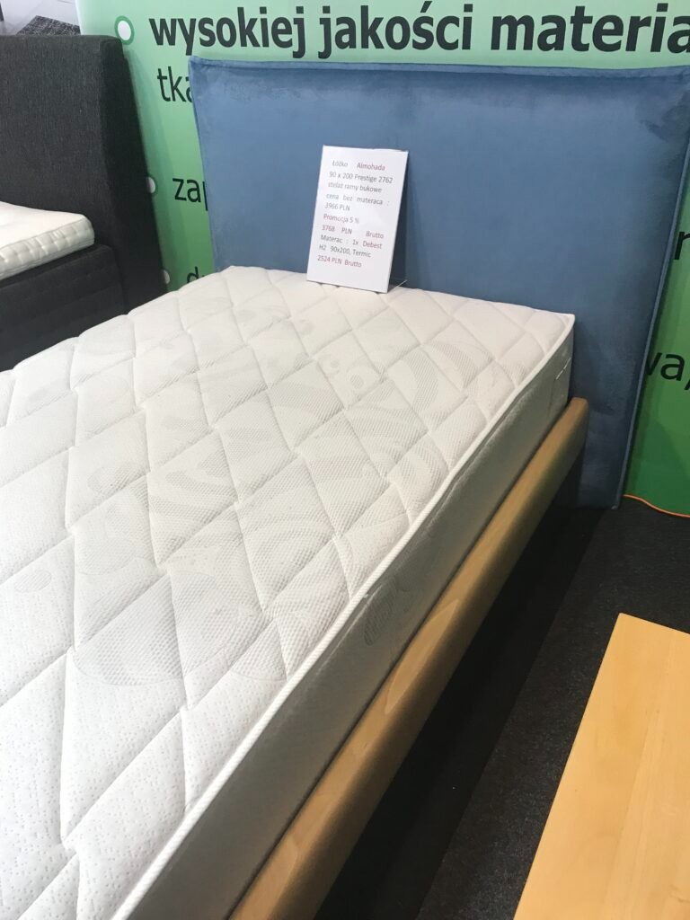 Łóżko z niebieskim tapicerowanym zagłówkiem i białym materacem w ramie z jasnego drewna, widoczna etykieta z ceną i specyfikacją.