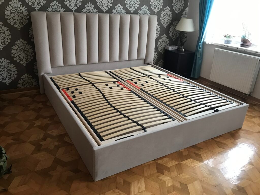 Rama łóżka tapicerowanego z wysokim wezgłowiem w odcieniach beżu, z drewnianym stelażem wewnątrz, ustawiona w pokoju z wzorzystą tapetą.