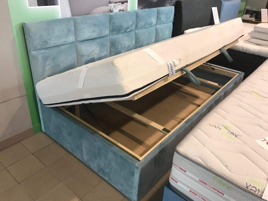 Łóżko tapicerowane z pojemnikiem na pościel w kolorze pastelowego błękitu, z podniesionym materacem ukazującym mechanizm otwierania i wnętrze skrzyni.