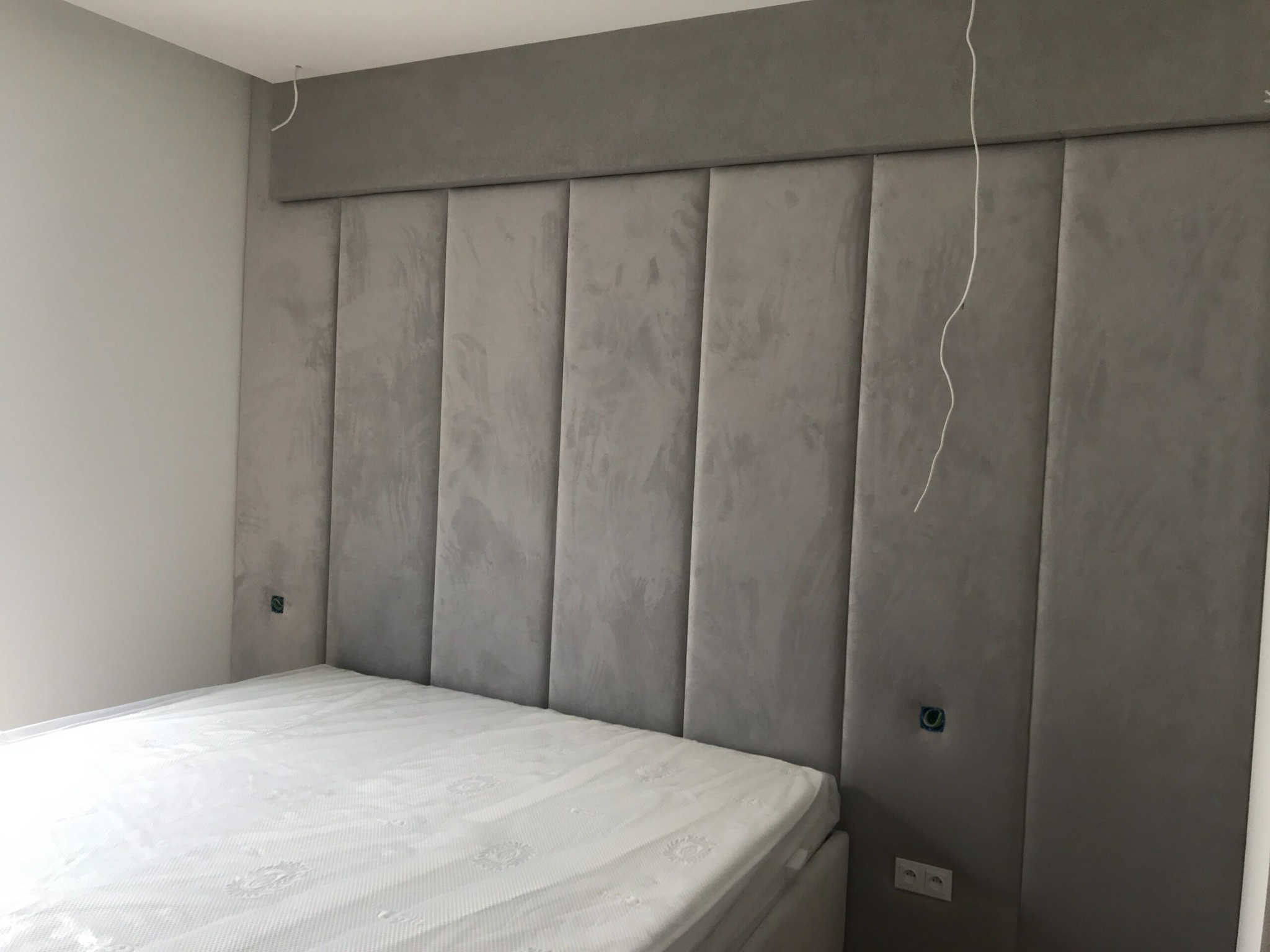 Szary tapicerowany zagłówek łóżka z panelami pionowymi, widoczne gniazdka elektryczne i wystające kable, łóżko z materacem w folii ochronnej.