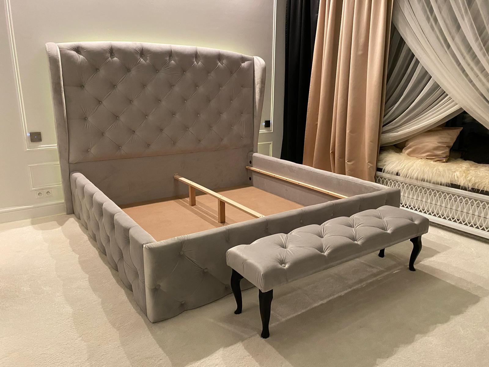 Szare, tapicerowane łóżko z wysokim wezgłowiem i pufą w stylu glamour, widoczne drewniane elementy konstrukcyjne, w tle zasłony i dekoracyjny szezlong.
