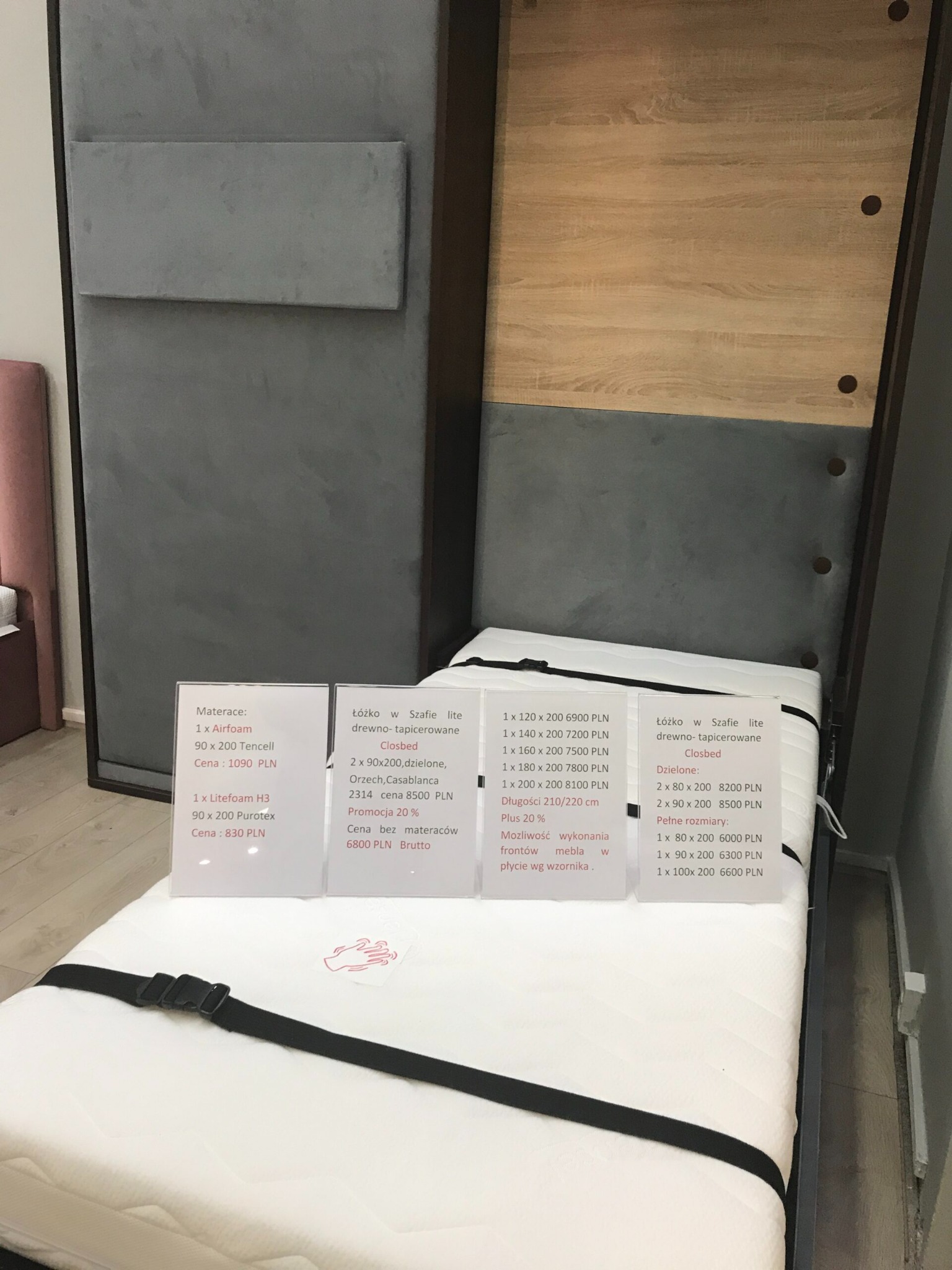 Łóżko chowane w szafie, w pozycji złożonej, z widocznym materacem zabezpieczonym pasami, obok widoczne kartki z cenami i specyfikacją materacy i łóżka.