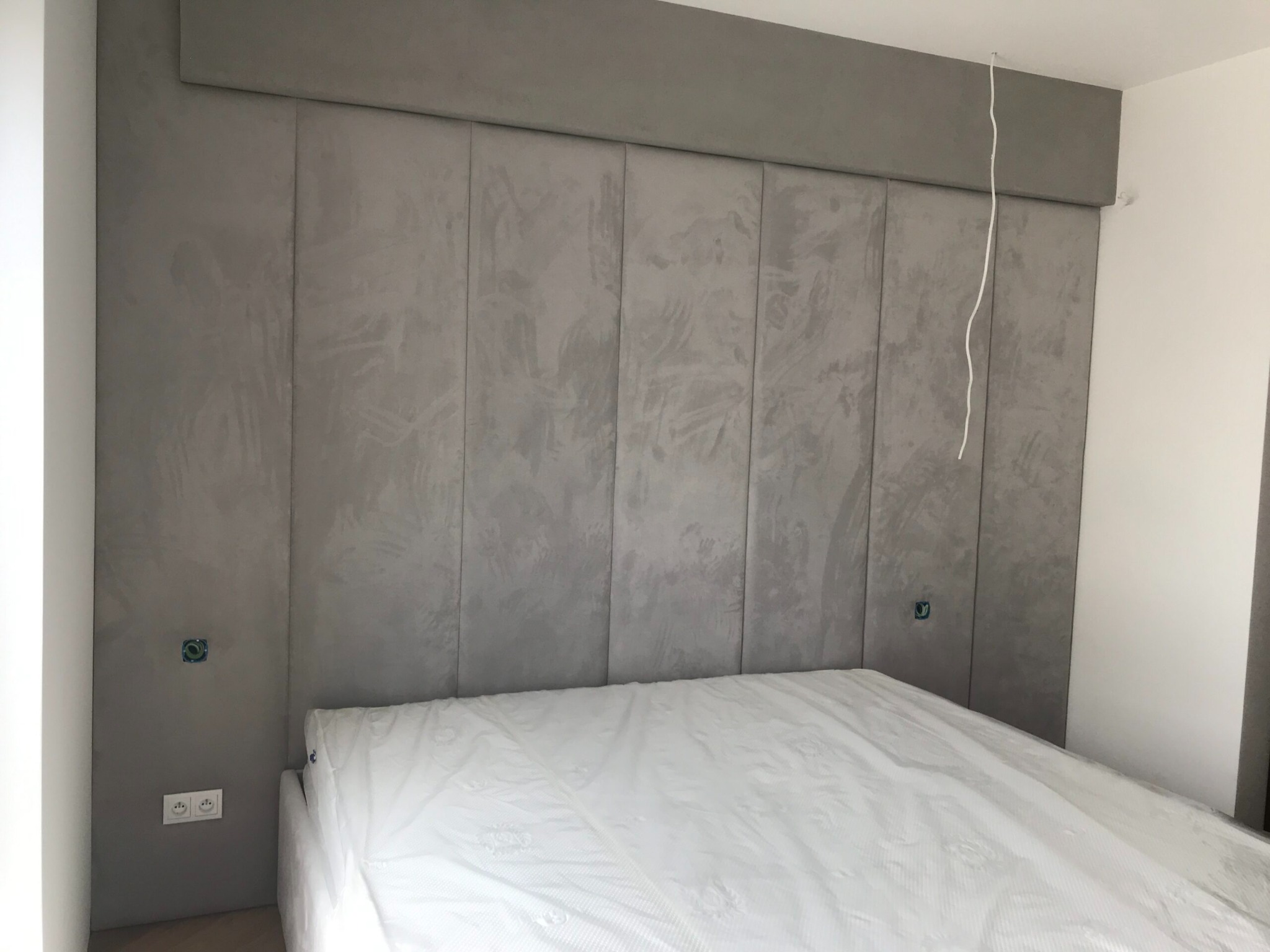 Szary, tapicerowany panel ścienny za łóżkiem z materacem w folii ochronnej, widoczne gniazdka elektryczne i wystające kable.