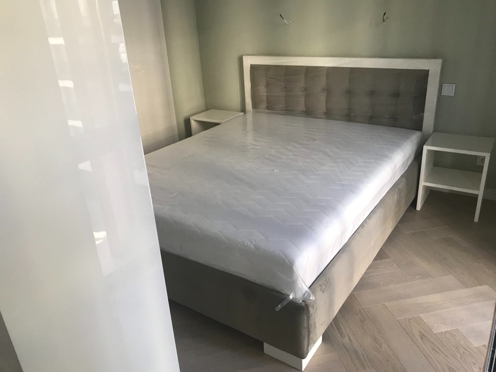 Nowe łóżko tapicerowane z szarą ramą i zagłówkiem, nadal owinięte w folię ochronną, ustawione w sypialni z drewnianą podłogą w jodełkę i białymi szafkami nocnymi.