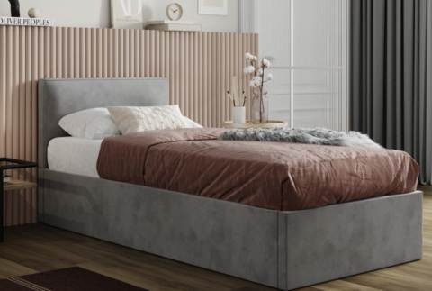 Szare łóżko tapicerowane z pojemnikiem na pościel, z wezgłowiem, nakryte brązową narzutą i dekoracyjną poduszką, na tle ściany z pionowych paneli i zasłon.