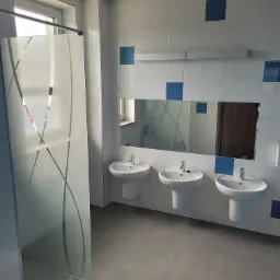 Publiczna łazienka z trzema białymi umywalkami, lustrem z oświetleniem i kabiną prysznicową z matowego szkła z wzorem.