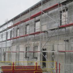 Spectom - Budynek w trakcie prac elewacyjnych, otoczony rusztowaniami z czerwonymi elementami. Widoczne okna i fragmenty ocieplenia. Na pierwszym planie barierki ochronne.