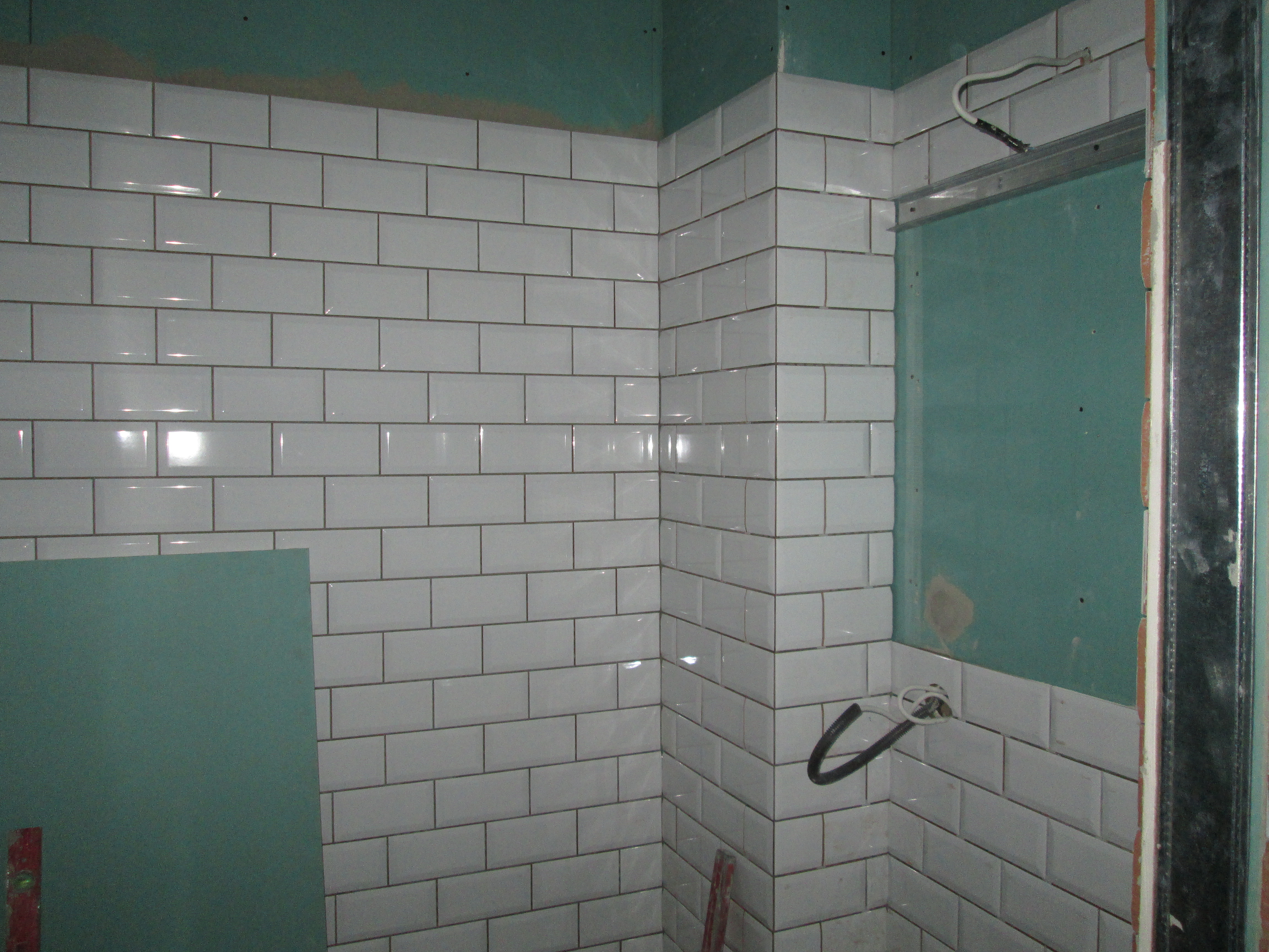 Białe, prostokątne płytki ułożone w cegiełkę na ścianie w rogu pomieszczenia. Widoczna płyta kartonowo-gipsowa i metalowy profil.