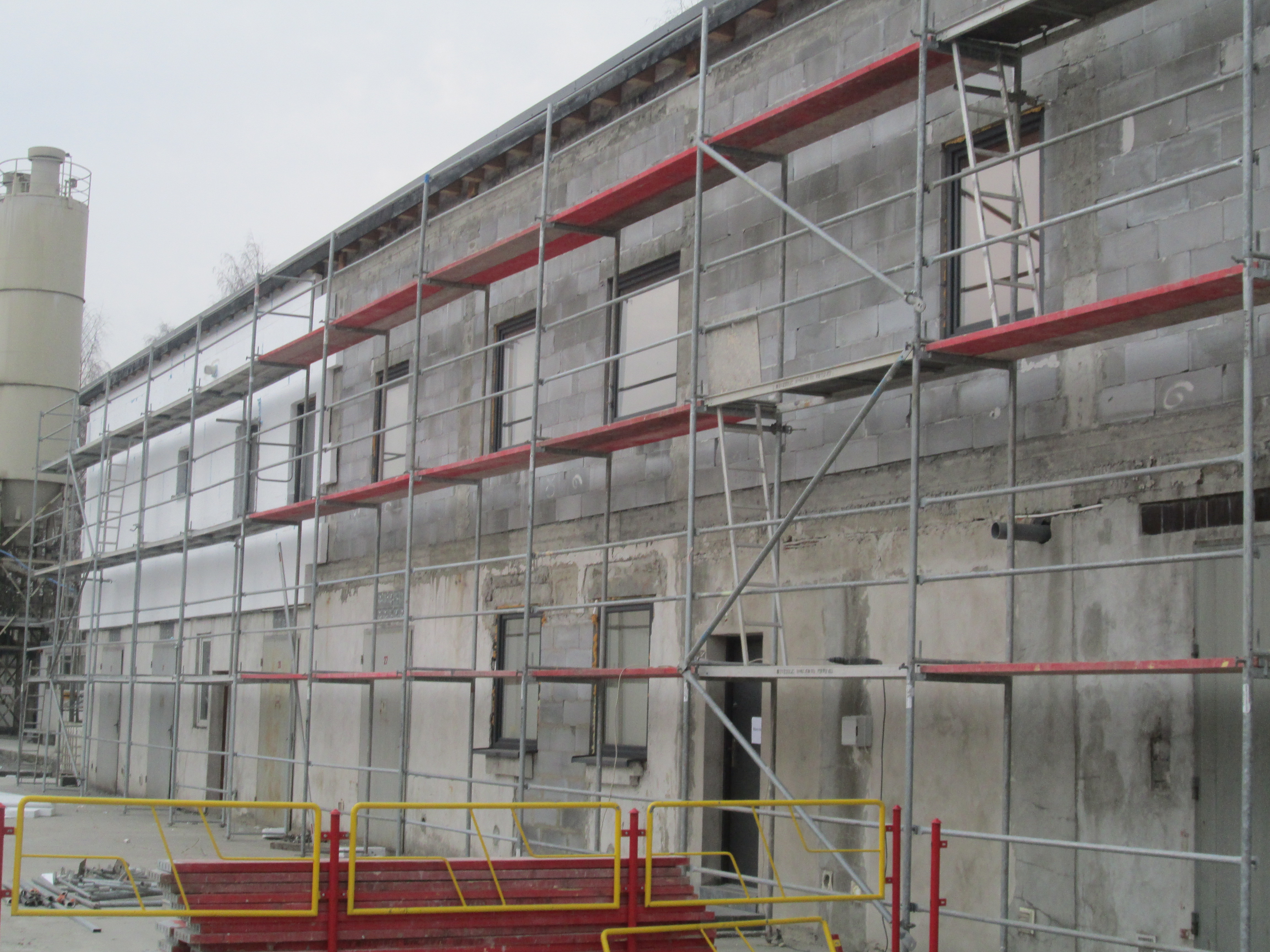 Budynek w trakcie prac elewacyjnych, otoczony rusztowaniami z czerwonymi elementami. Widoczne okna i fragmenty ocieplenia. Na pierwszym planie barierki ochronne.