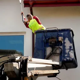 Pracownik w podnośniku czyści rynny myjką ciśnieniową w Łodzi. Czerwona dachówka i biała elewacja budynku w tle. Żółta kamizelka ochronna.