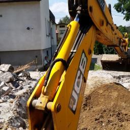 Vikop - Żółta koparka JCB podczas prac ziemnych, z gruzem i stertą ziemi na pierwszym planie, operator koparki siedzący w tle przy budynku.