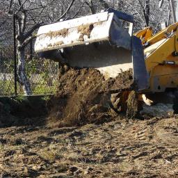 Vikop - Żółta koparko-ładowarka JCB 3CX z pełną łyżką ziemi w trakcie prac ziemnych na terenie budowy z widoczną siatką ogrodzeniową w tle.