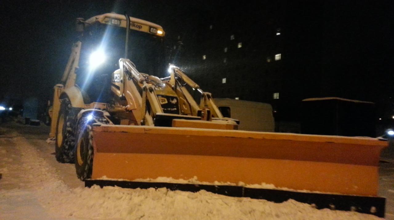 Żółta spycharka do śniegu z włączonymi światłami, stojąca na ośnieżonym terenie w nocy, widoczne budynki w tle.