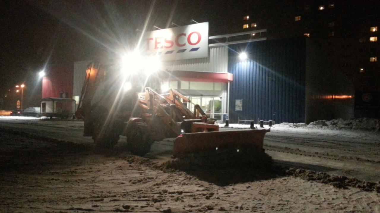 Pomarańczowy traktor z pługiem odśnieża parking przed sklepem Tesco w nocy, oświetlony reflektorami.