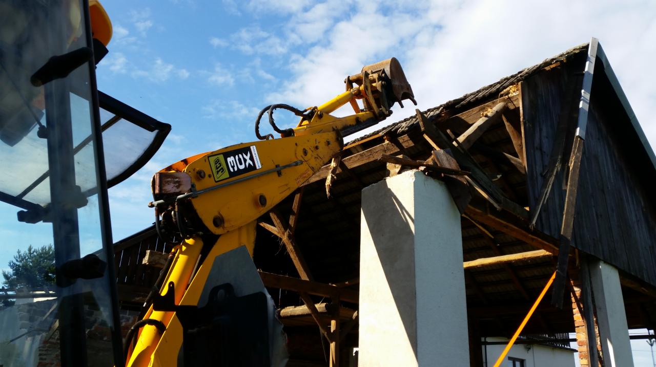Żółta koparka JCB demontuje drewniany dach starego budynku z białymi kolumnami w słoneczny dzień.