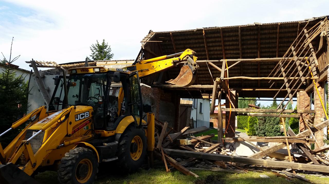 Żółta koparko-ładowarka JCB podczas rozbiórki starej, częściowo zawalonej stodoły z ceglanymi ścianami i drewnianą konstrukcją dachu.