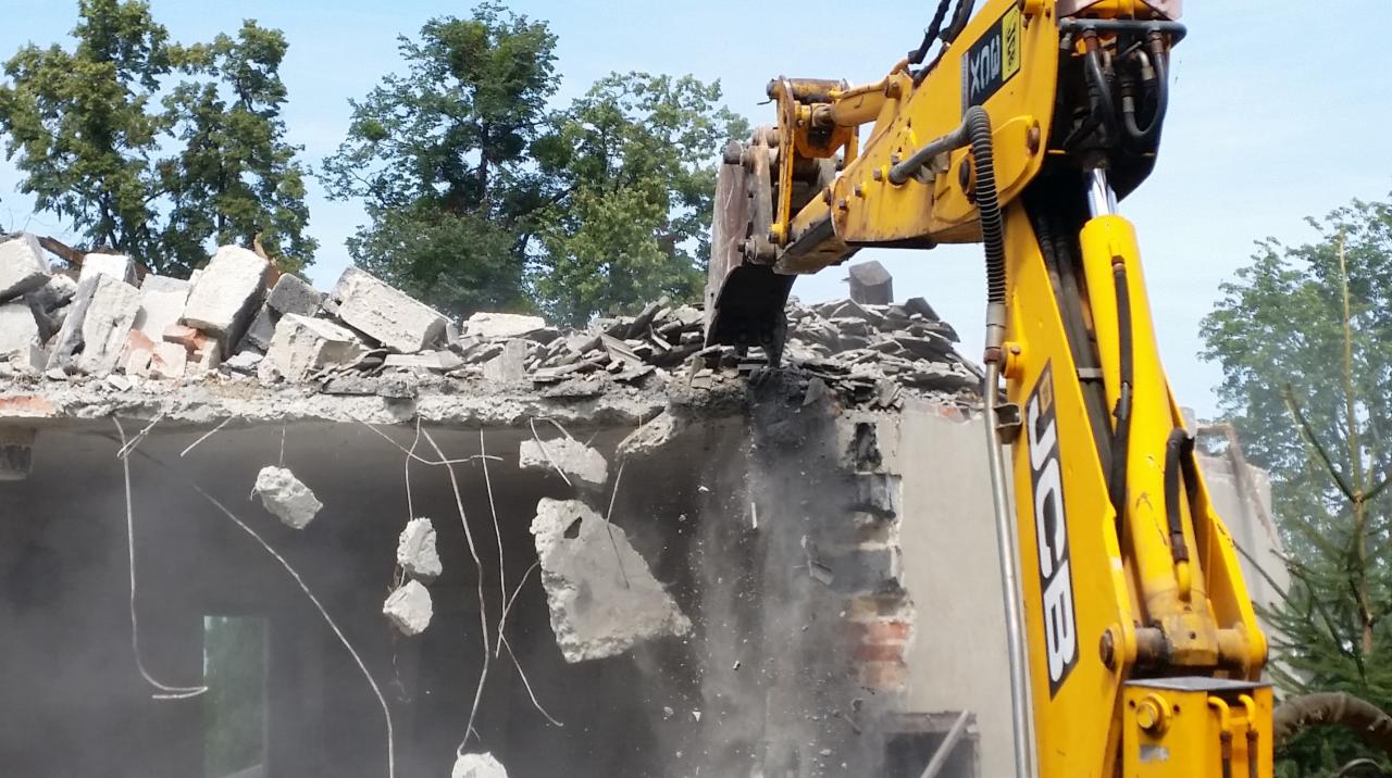 Żółta koparka JCB kruszy resztki betonowej konstrukcji budynku, w tle drzewa i niebieskie niebo.
