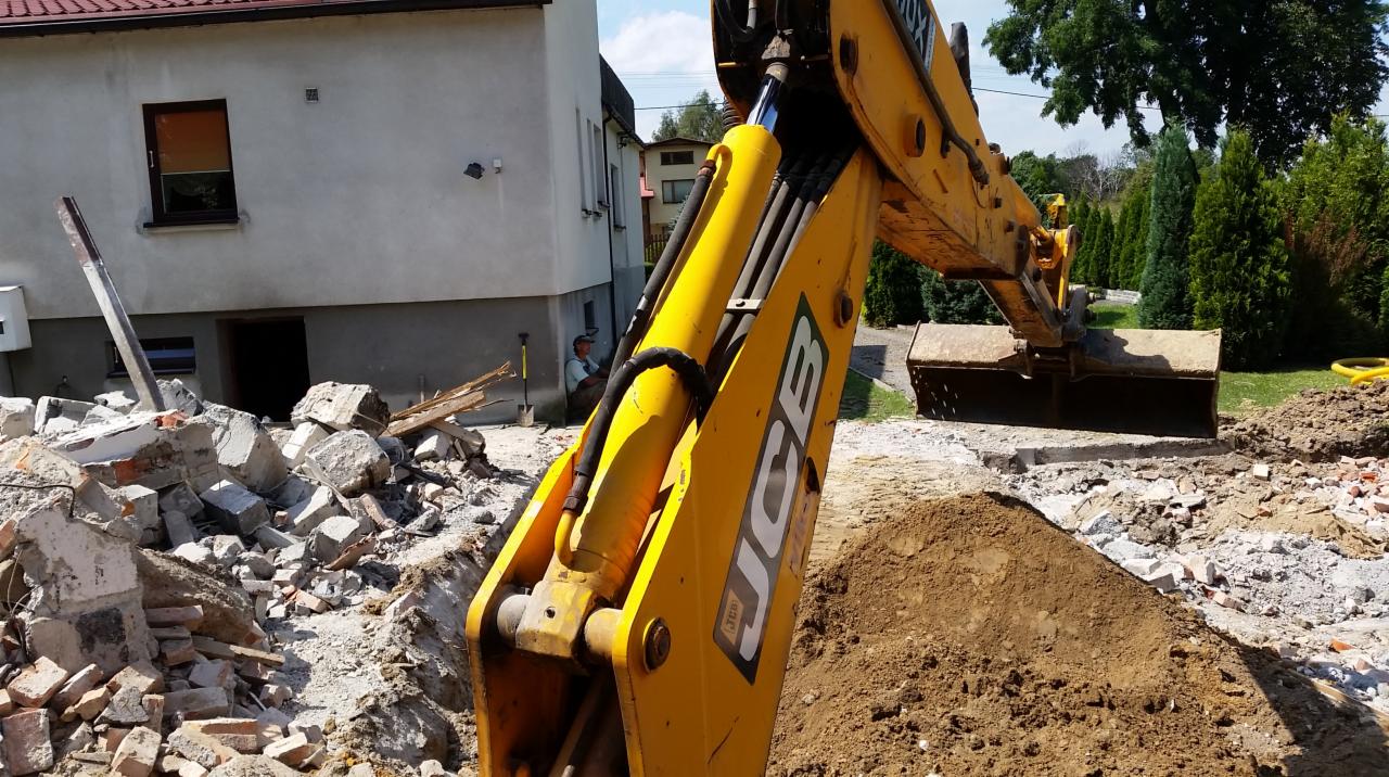 Żółta koparka JCB podczas prac ziemnych, z gruzem i stertą ziemi na pierwszym planie, operator koparki siedzący w tle przy budynku.