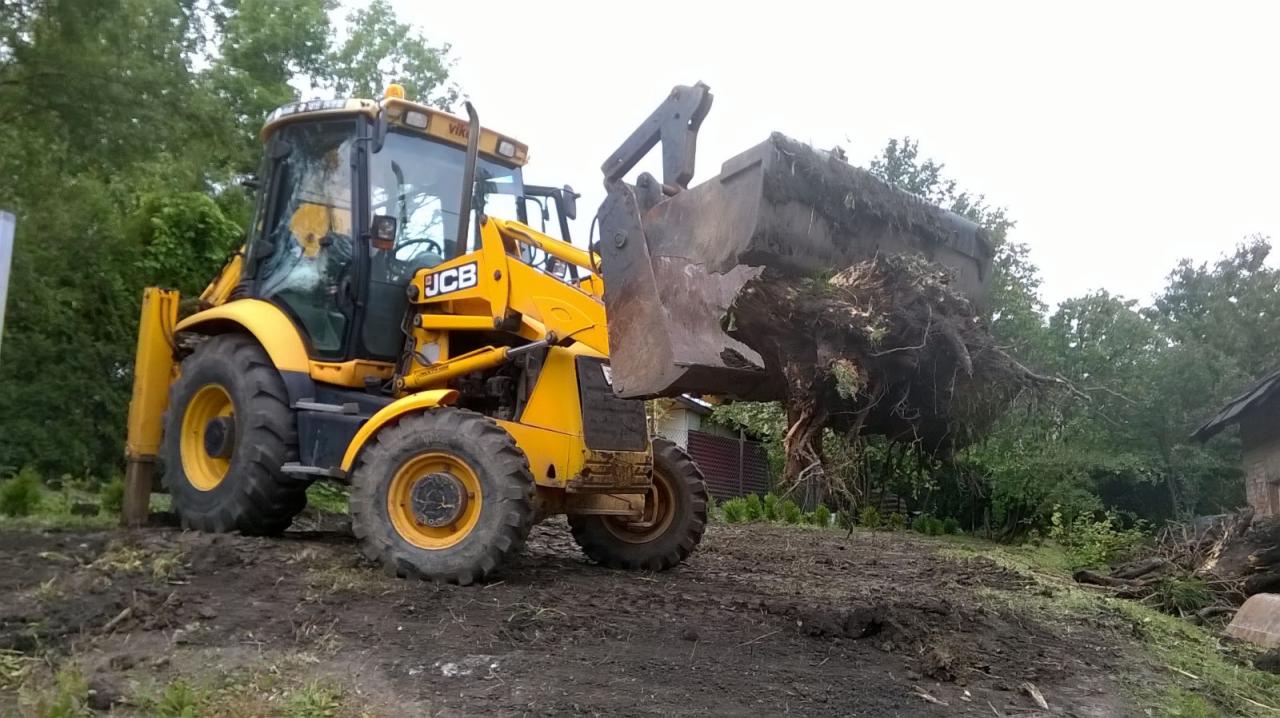 Żółta koparko-ładowarka JCB z pełną łyżką korzeni drzew na nierównym, błotnistym terenie, w tle zieleń i fragment budynku.