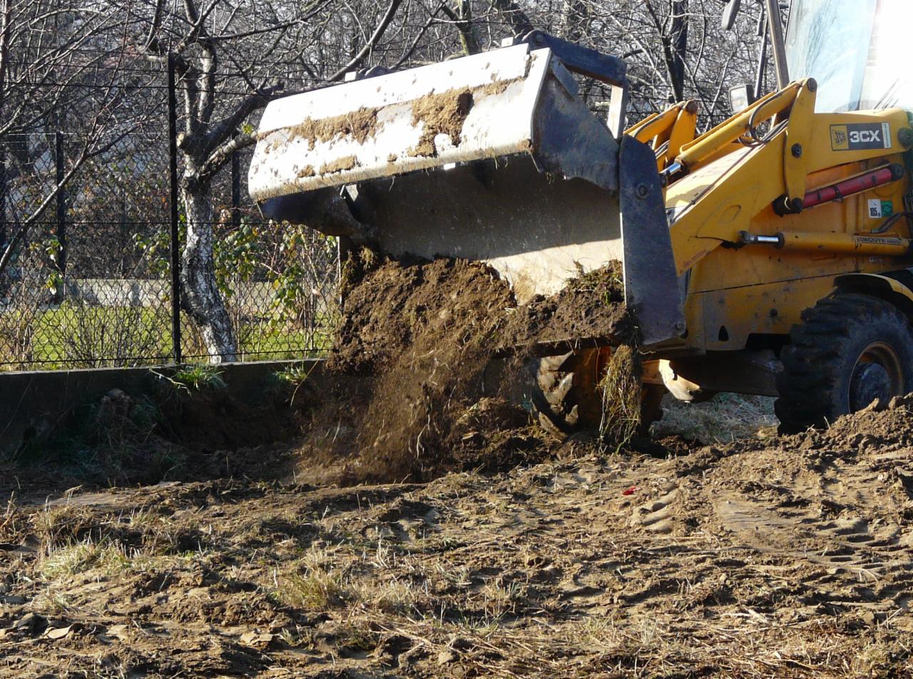 Żółta koparko-ładowarka JCB 3CX z pełną łyżką ziemi w trakcie prac ziemnych na terenie budowy z widoczną siatką ogrodzeniową w tle.