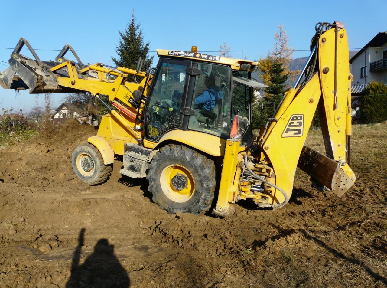 Żółta koparko-ładowarka JCB na nierównym, błotnistym terenie, widoczny operator w kabinie, drzewa w tle, cień osoby robiącej zdjęcie na ziemi.