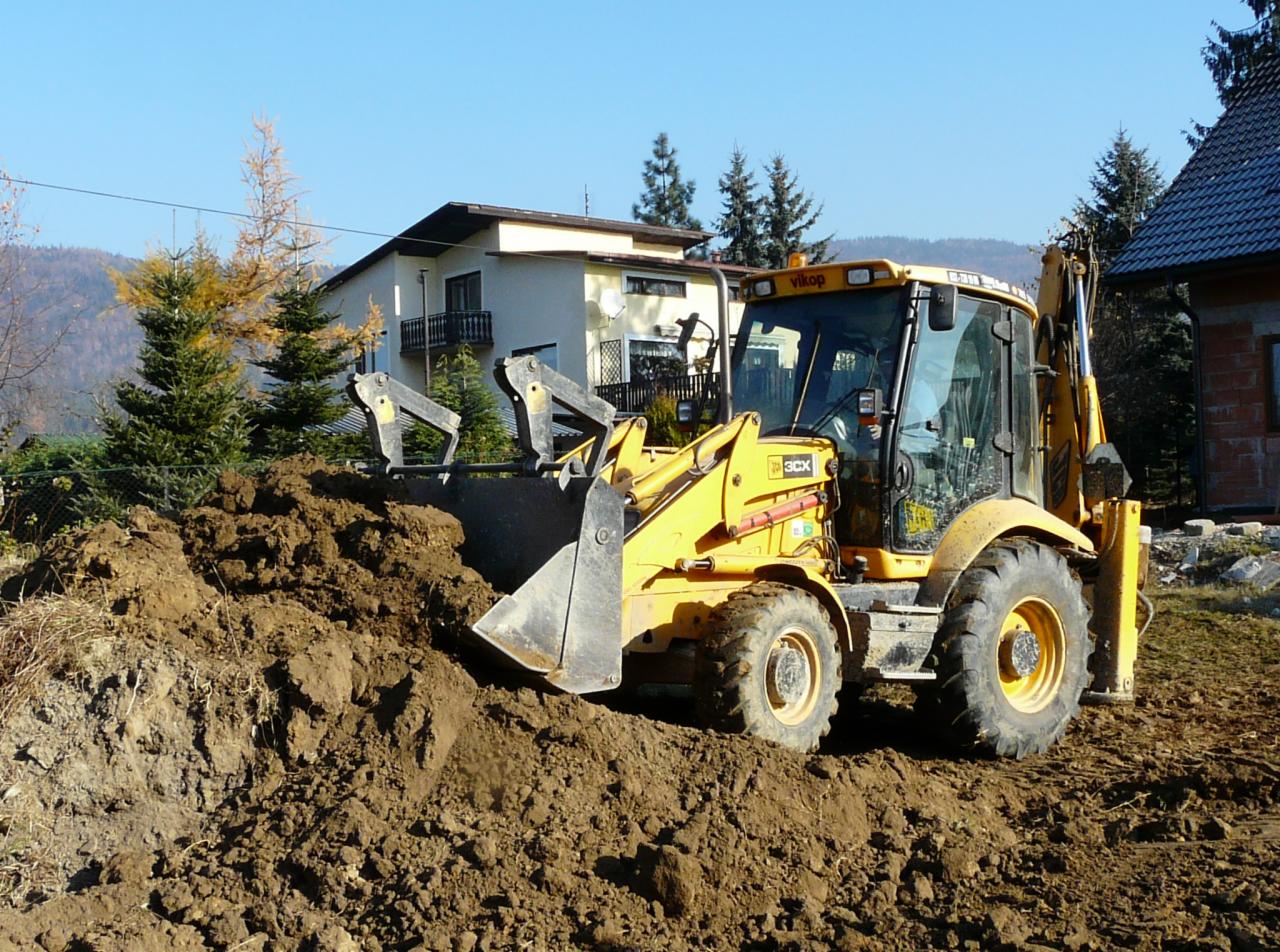 Żółta koparko-ładowarka Vikop 3CX nabiera ziemię na tle domu jednorodzinnego i drzew