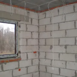 Surowy mur z oknem i wyprowadzonymi przewodami elektrycznymi. Widoczne puszki instalacyjne i kable zwisające ze ściany i sufitu, etap prac w nowym budynku.