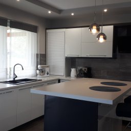 Usługi stolarsko budowlane Józef Grapa - Nowoczesna kuchnia z białymi, błyszczącymi frontami, czarnym zlewem pod oknem z roletami rzymskimi, wyspą z blatem w kolorze frontów i czarną podstawą, oraz wiszącymi lampami z przydymionego szkła.