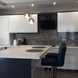 Usługi stolarsko budowlane Józef Grapa - Nowoczesna kuchnia z wyspą, białymi frontami szafek, szarym blatem i barkowymi krzesłami z ciemnego weluru, oświetlona wiszącymi lampami kulami.