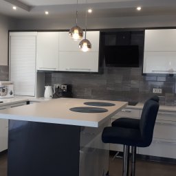 Usługi stolarsko budowlane Józef Grapa - Nowoczesna kuchnia z wyspą i dwoma hokerami w odcieniach bieli i szarości, z wiszącymi lampami kulami nad blatem, ciemnoszarym backsplash z płytek i sprzętami AGD.