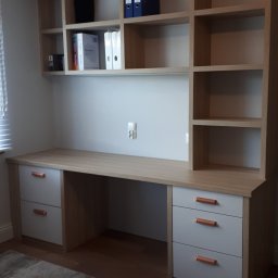 Usługi stolarsko budowlane Józef Grapa - Jasny biurko z szufladami i regałem na książki, z szarymi frontami i miedzianymi uchwytami, uzupełnione o segregatory i książki, ustawione na tle jasnej ściany z gniazdkiem.