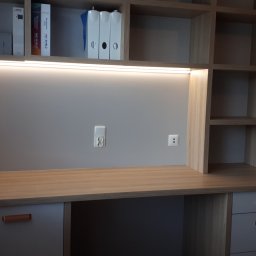 Usługi stolarsko budowlane Józef Grapa - Biurko z szufladami i nadstawką z półkami, oświetlone listwą LED, z widocznymi gniazdkami elektrycznymi na ścianie, obok okna z żaluzjami.