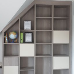 Usługi stolarsko budowlane Józef Grapa - Zabudowa wnęki pod skosem z półkami i szafkami w odcieniach szarości i bieli, na półkach książki, globus, segregatory i ramki z certyfikatami.