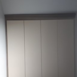 Usługi stolarsko budowlane Józef Grapa - Czterodrzwiowa szafa wnękowa z jasnymi frontami i szarobrązową ramą, dopasowana do skosu sufitu, w pomieszczeniu z jasną podłogą.