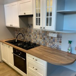 Usługi stolarsko budowlane Józef Grapa - Nowoczesna, biała kuchnia z drewnianym blatem, czarnym zlewem i płytą grzewczą, zdobiona mozaiką nad blatem, z otwartymi półkami i częściowo widoczną ręką osoby sprzątającej.