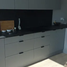 Szare, minimalistyczne szafki kuchenne z czarnym blatem i panelami ściennymi, na blacie deska do krojenia, miseczki i przyprawy, w tle owoce w koszyku.