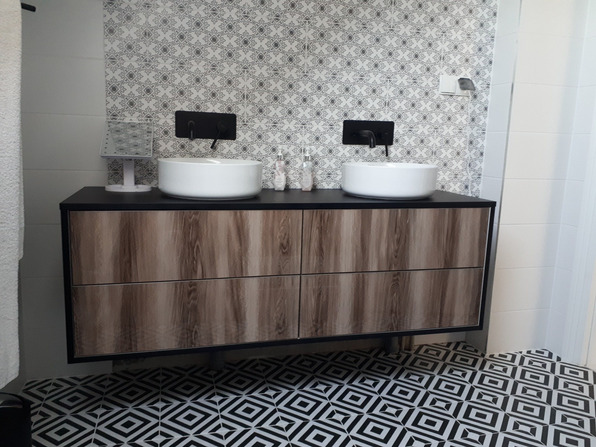 Łazienka z dwiema białymi umywalkami nablatowymi i czarną armaturą, szafka pod umywalkę z drewnopodobnym frontem, wzorzysta tapeta i podłoga w geometryczne wzory.