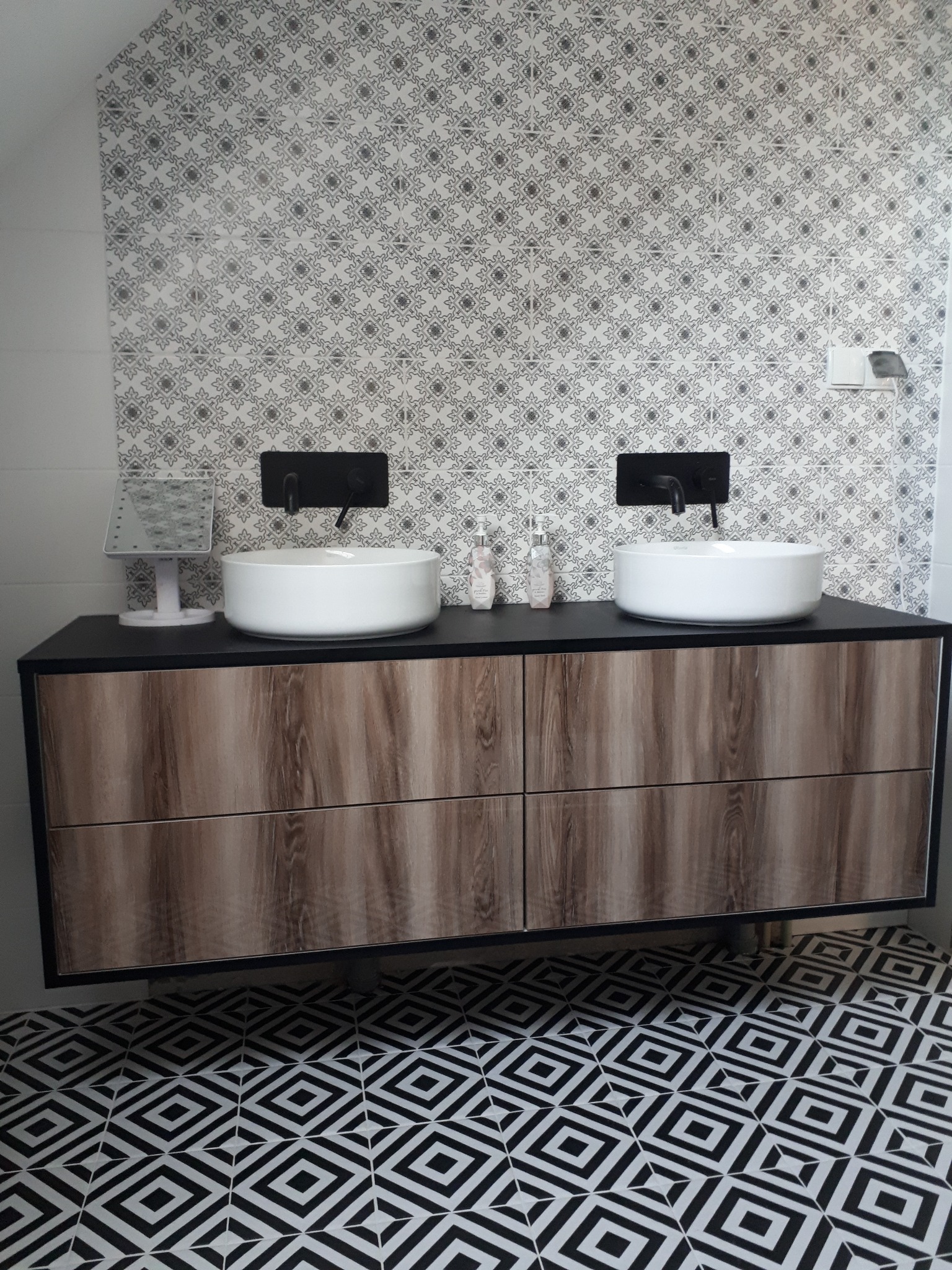Nowoczesna łazienka z wiszącą szafką pod umywalki nablatowe, czarną armaturą i wzorzystą tapetą oraz podłogą w geometryczny deseń.
