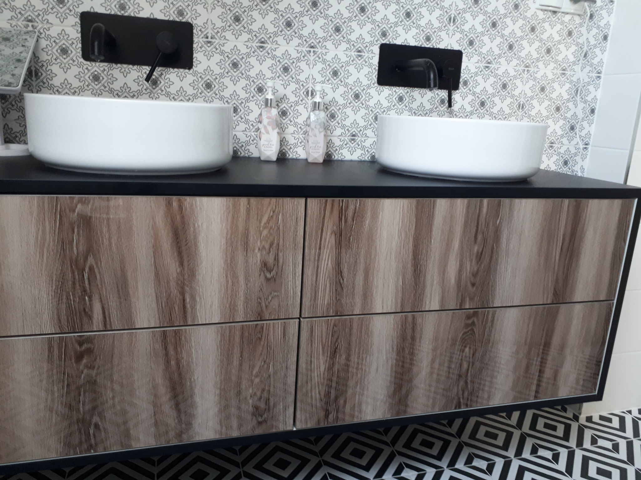 Łazienka z dwoma białymi umywalkami nablatowymi, czarną armaturą, szafką z frontami imitującymi drewno i wzorzystymi płytkami na ścianie i podłodze.