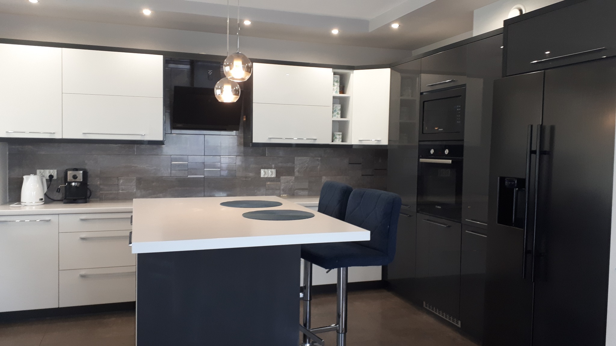 Nowoczesna kuchnia z białymi i grafitowymi szafkami, wyspą z białym blatem i dwoma granatowymi krzesłami barowymi, oświetlona wiszącymi lampami kulami.