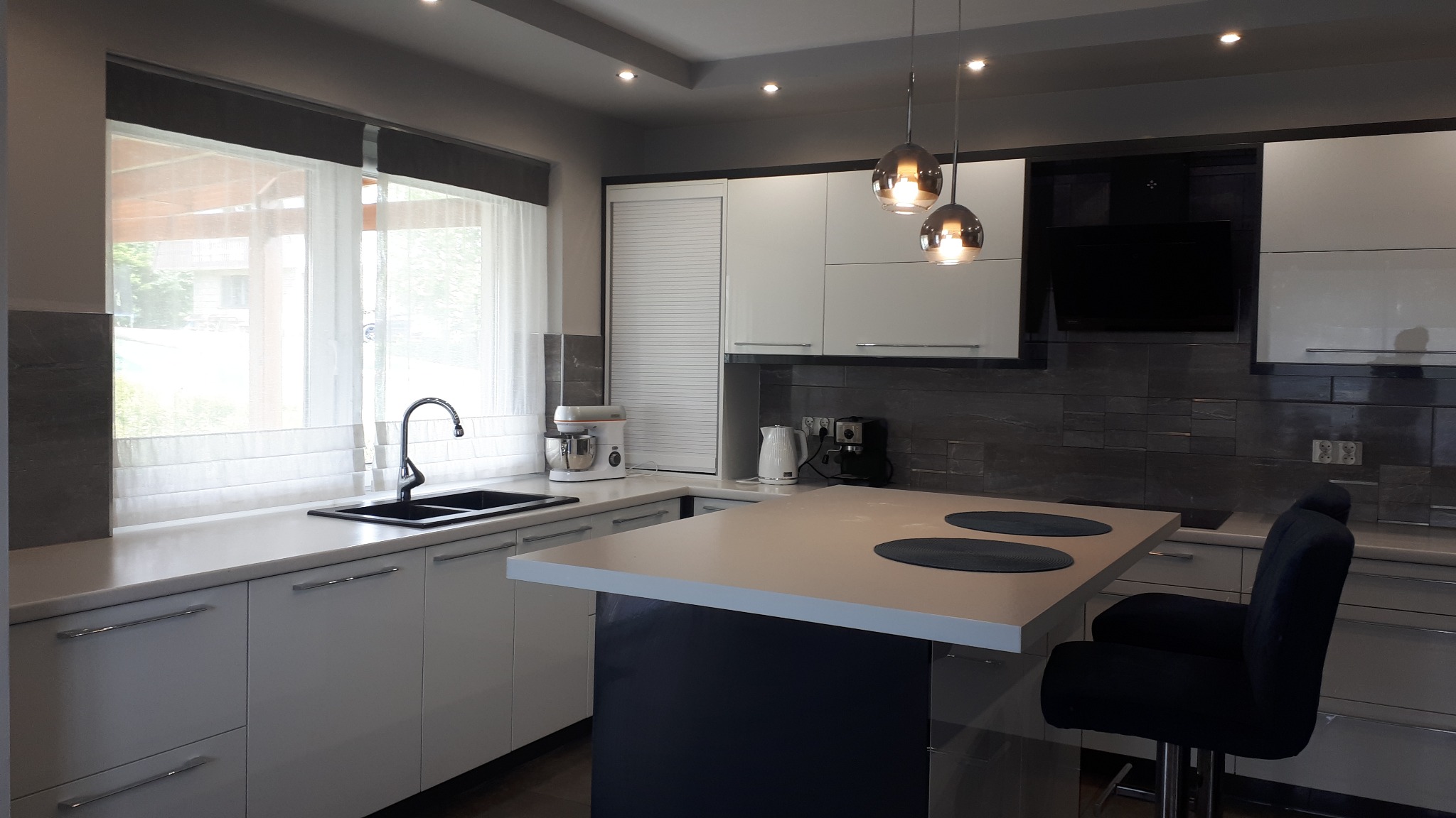 Nowoczesna kuchnia z białymi, błyszczącymi frontami, czarnym zlewem pod oknem z roletami rzymskimi, wyspą z blatem w kolorze frontów i czarną podstawą, oraz wiszącymi lampami z przydymionego szkła.