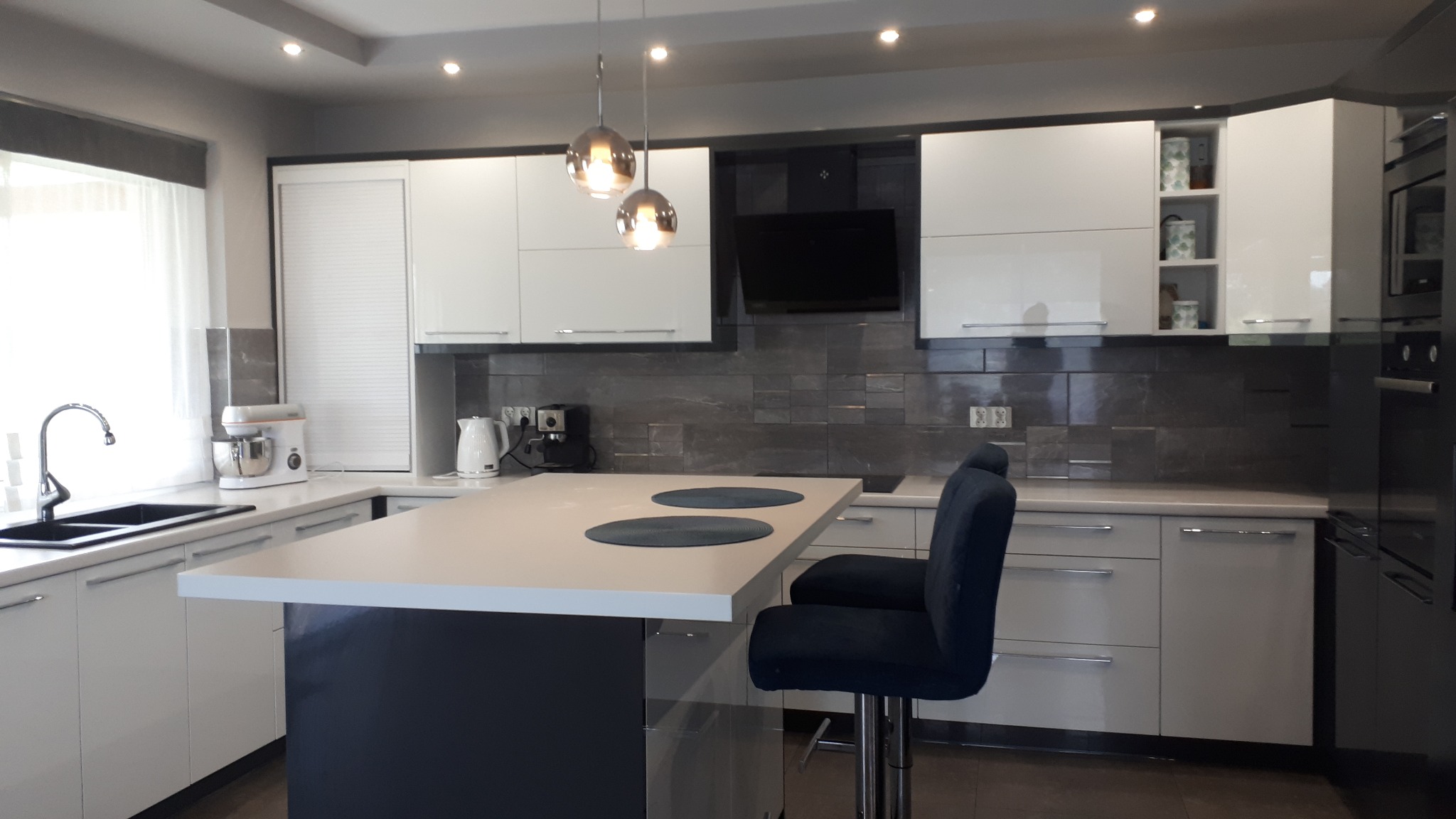 Nowoczesna kuchnia z wyspą, białymi frontami szafek, szarym blatem i barkowymi krzesłami z ciemnego weluru, oświetlona wiszącymi lampami kulami.