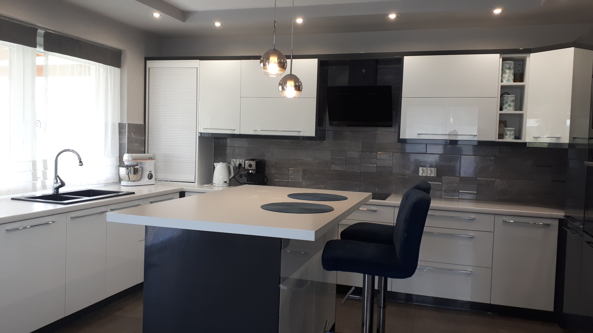Nowoczesna kuchnia z wyspą i dwoma hokerami w odcieniach bieli i szarości, z wiszącymi lampami kulami nad blatem, ciemnoszarym backsplash z płytek i sprzętami AGD.