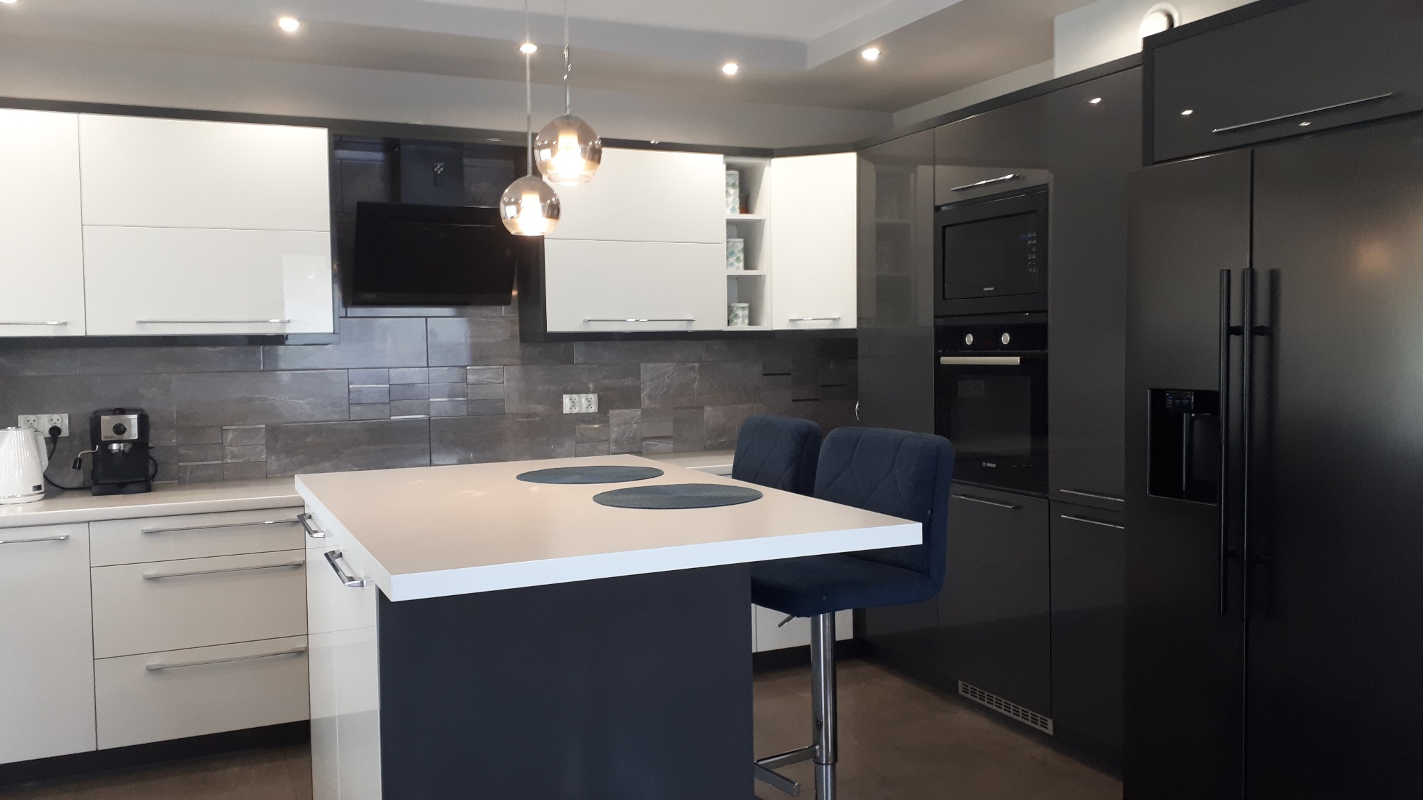Nowoczesna kuchnia z wyspą, biało-czarne szafki z połyskiem, szare płytki na ścianie, dwie niebieskie aksamitne hokery i szklane lampy wiszące.